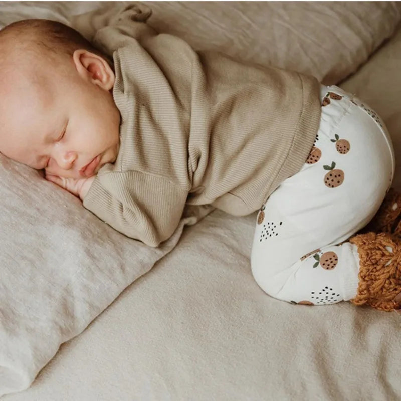 Slapende baby in beige trui en legging van biologisch katoen met natuurprint, op zacht beddengoed